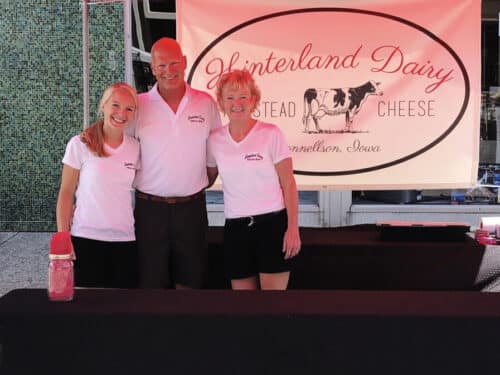 Three people standing by banquet tables at an outdoor farmers market with a Hinterland Dairy banner in the background